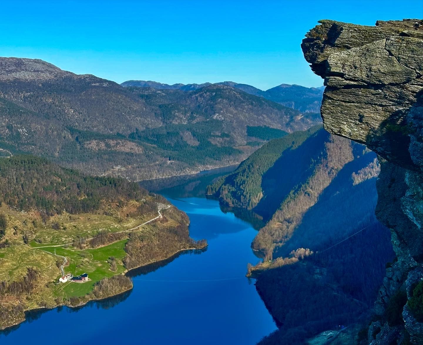 Jagged rock cliff overlooking a blue fjord winding through forested mountains with a small farm.