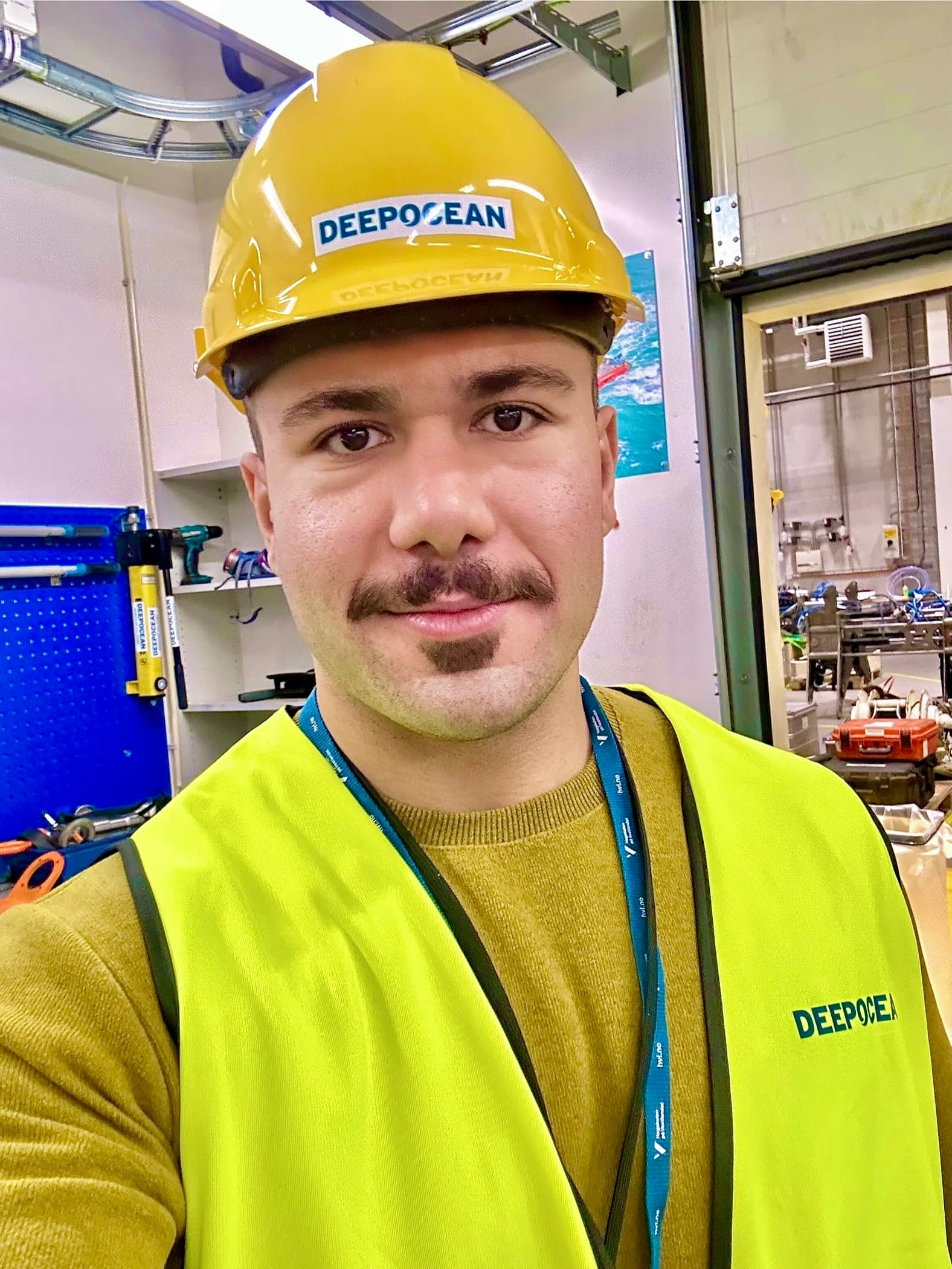 Man with a mustache wearing a yellow DeepOcean hard hat and safety vest in workshop.