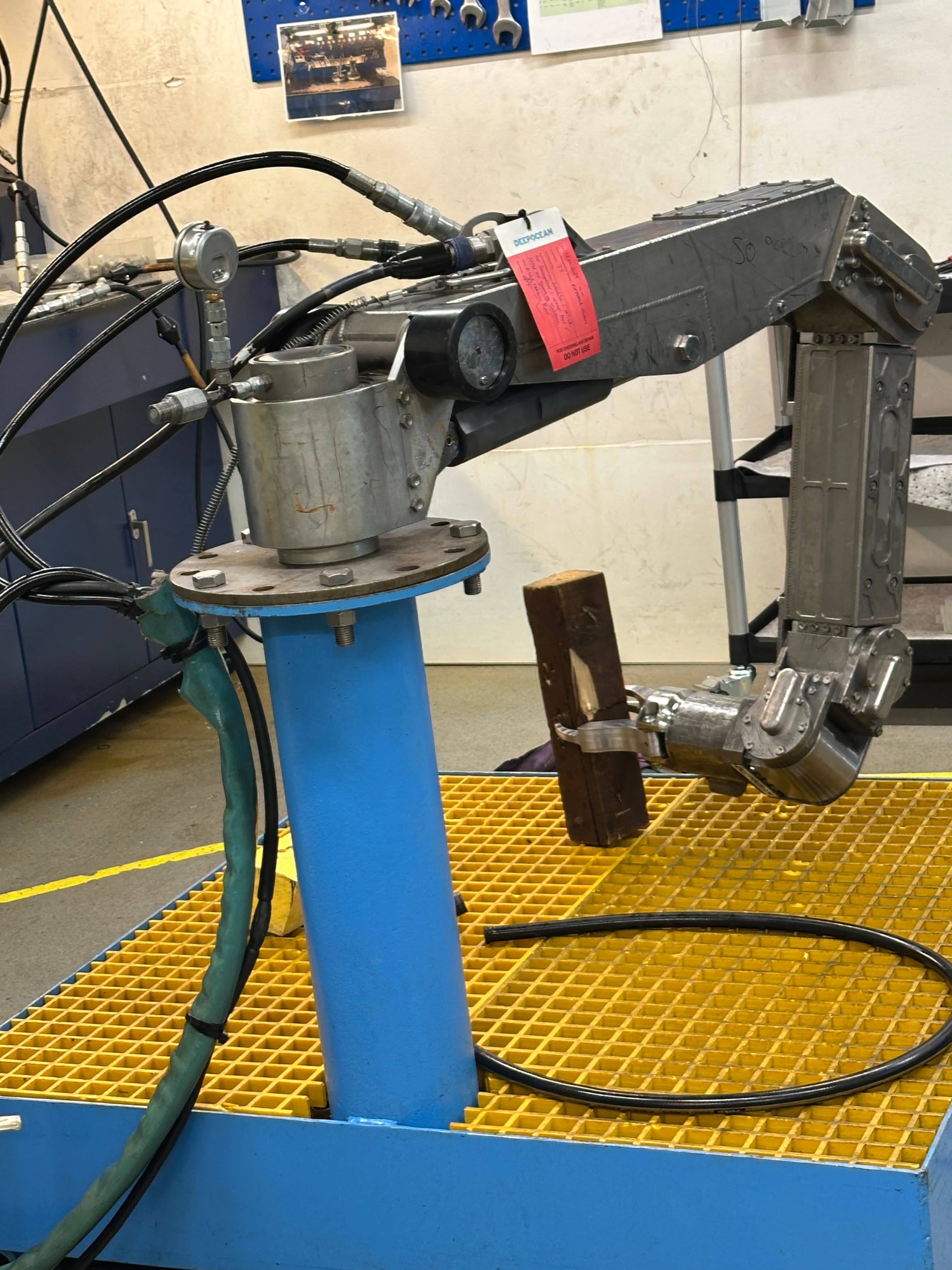 Silver robotic arm with hydraulic hoses mounted on a blue base over yellow floor grating.