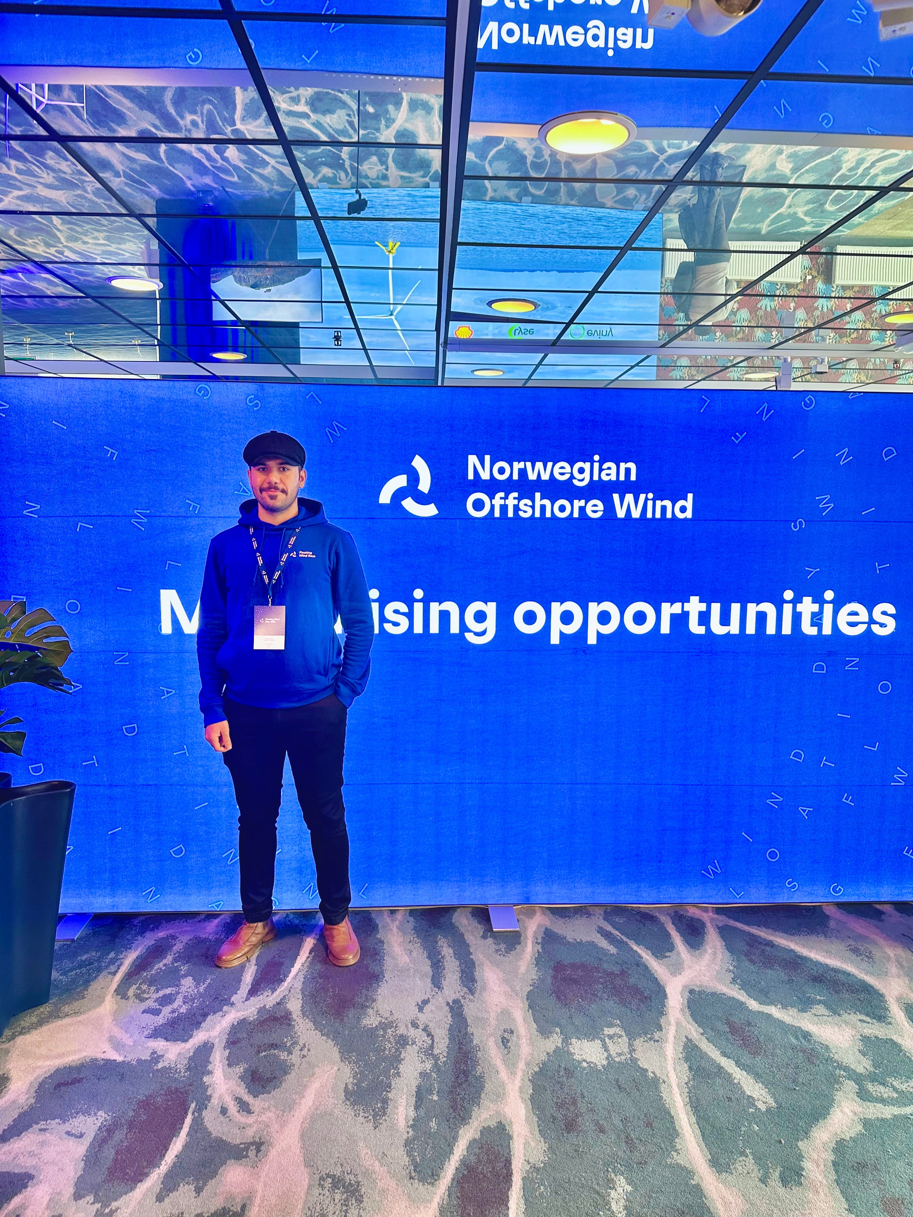 Man in blue hoodie stands before a blue Norwegian Offshore Wind event backdrop.