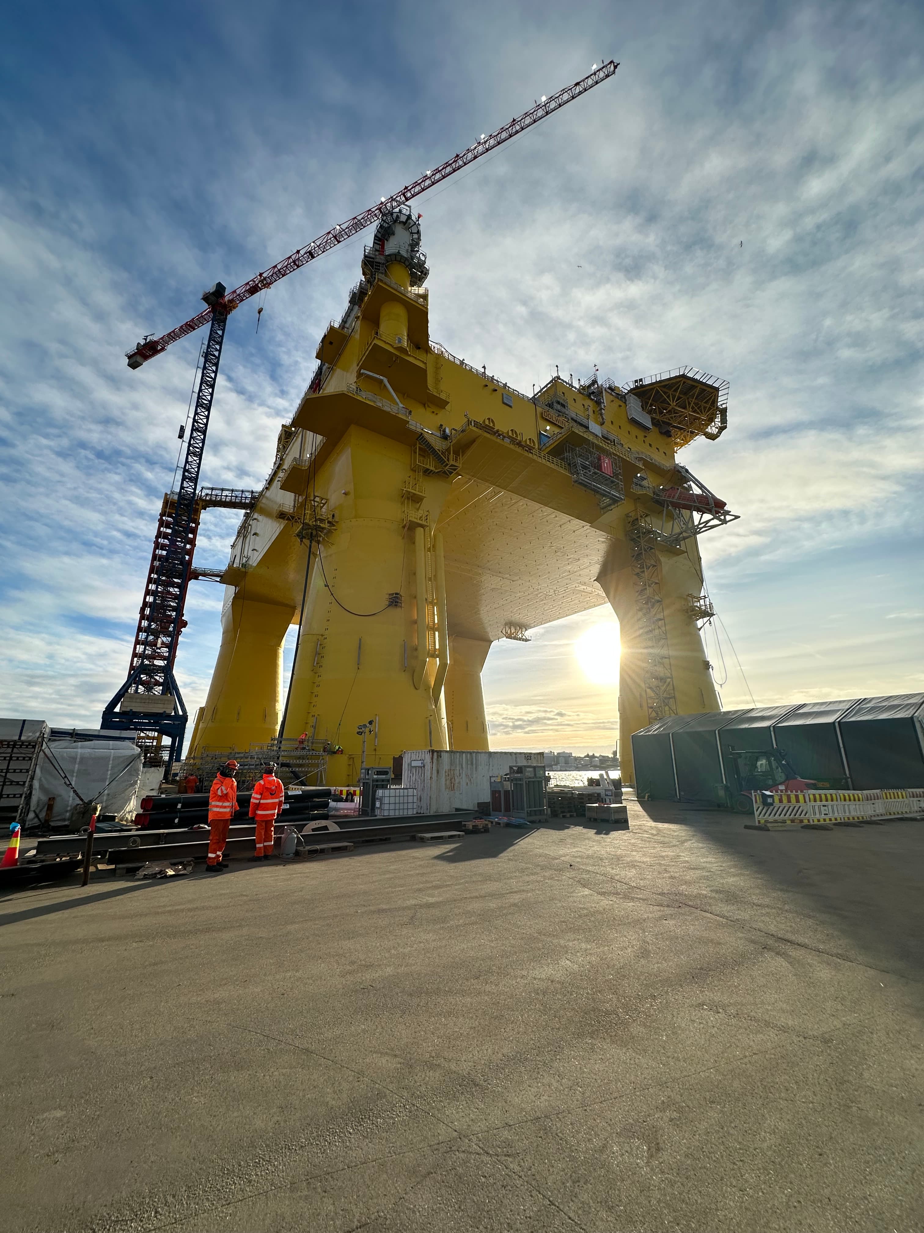 Towering yellow offshore platform at a shipyard with workers and a crane in bright sunlight.