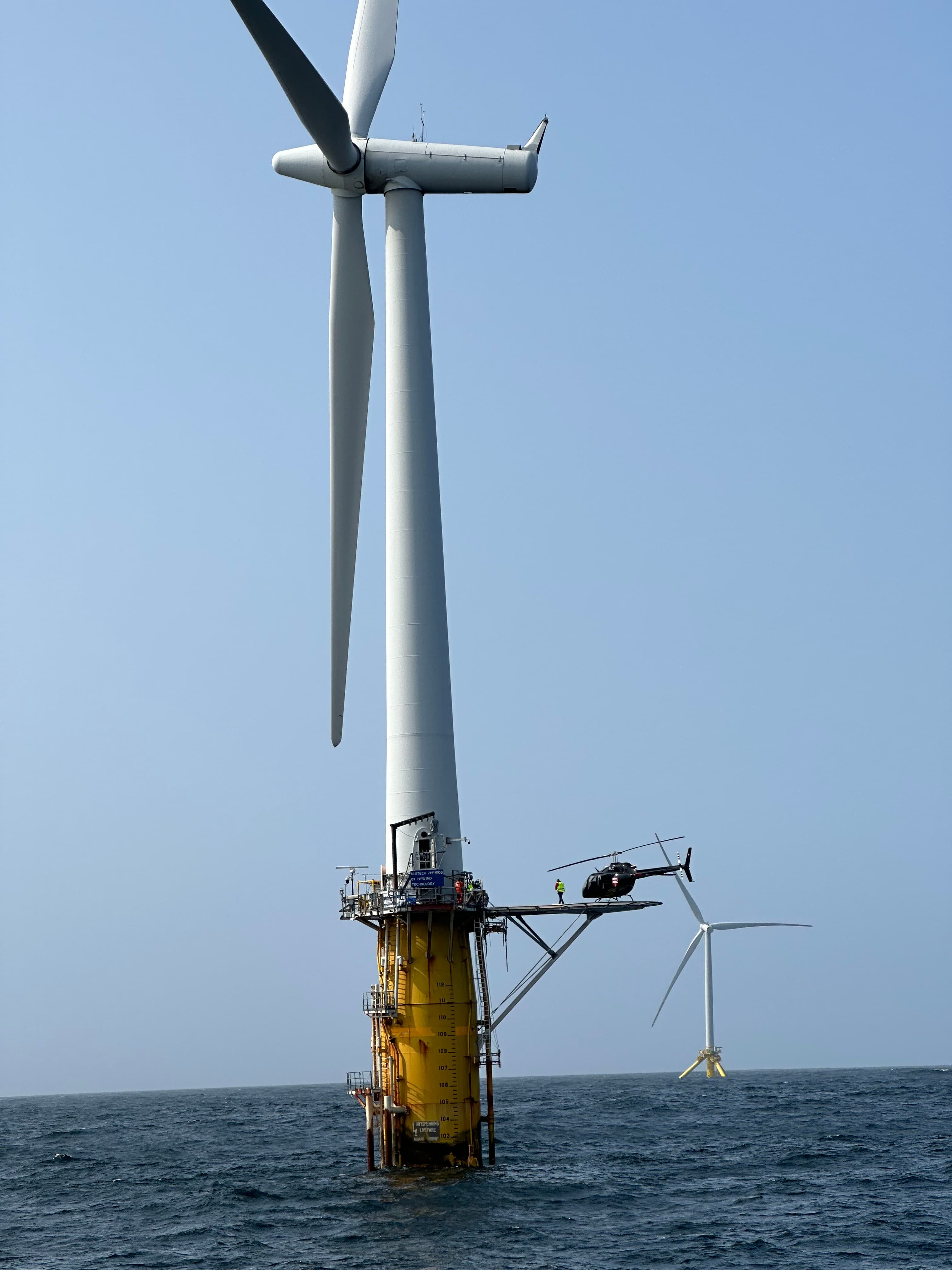 Black helicopter on a landing platform of a massive offshore wind turbine in the ocean.