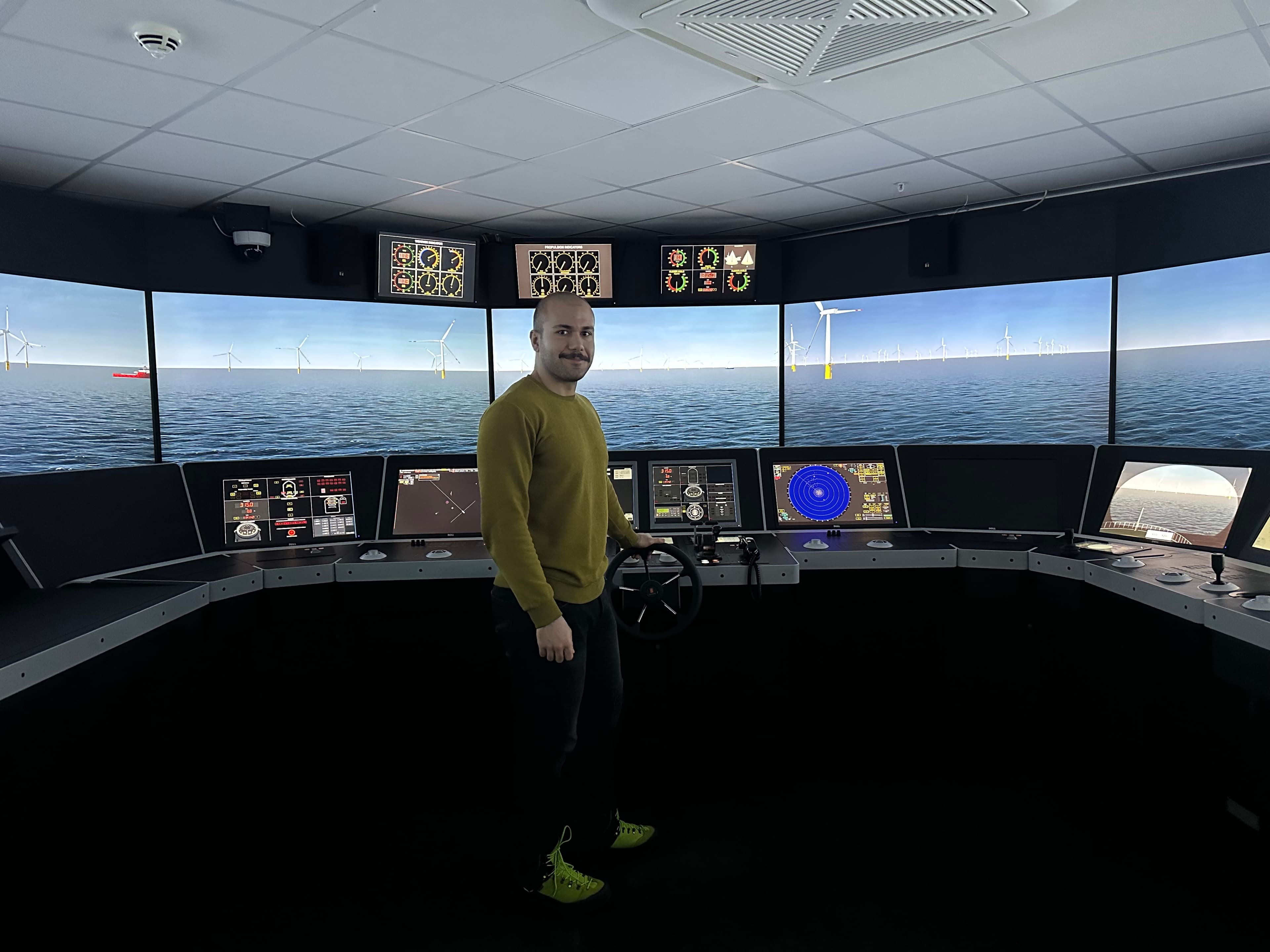 Man at a ship bridge simulator helm with panoramic screens showing offshore wind turbines.