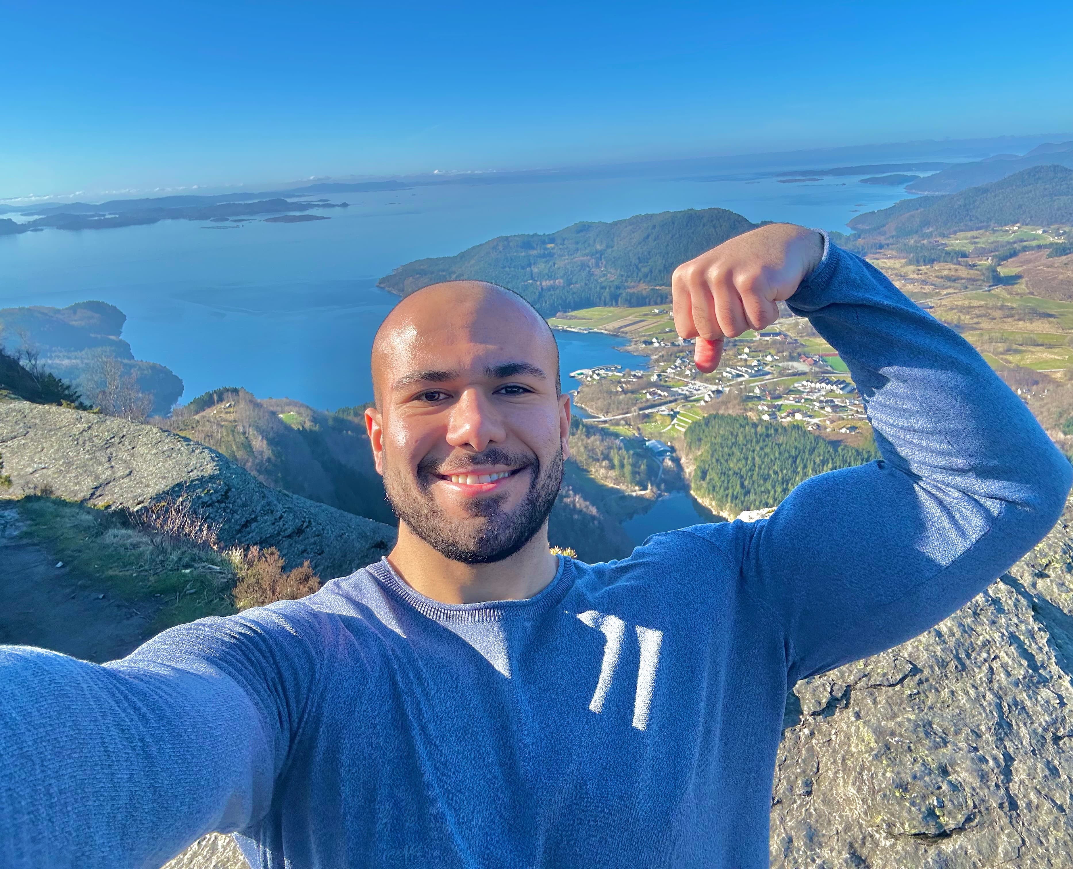 Smiling man flexing his bicep on a mountain peak overlooking a scenic fjord landscape.