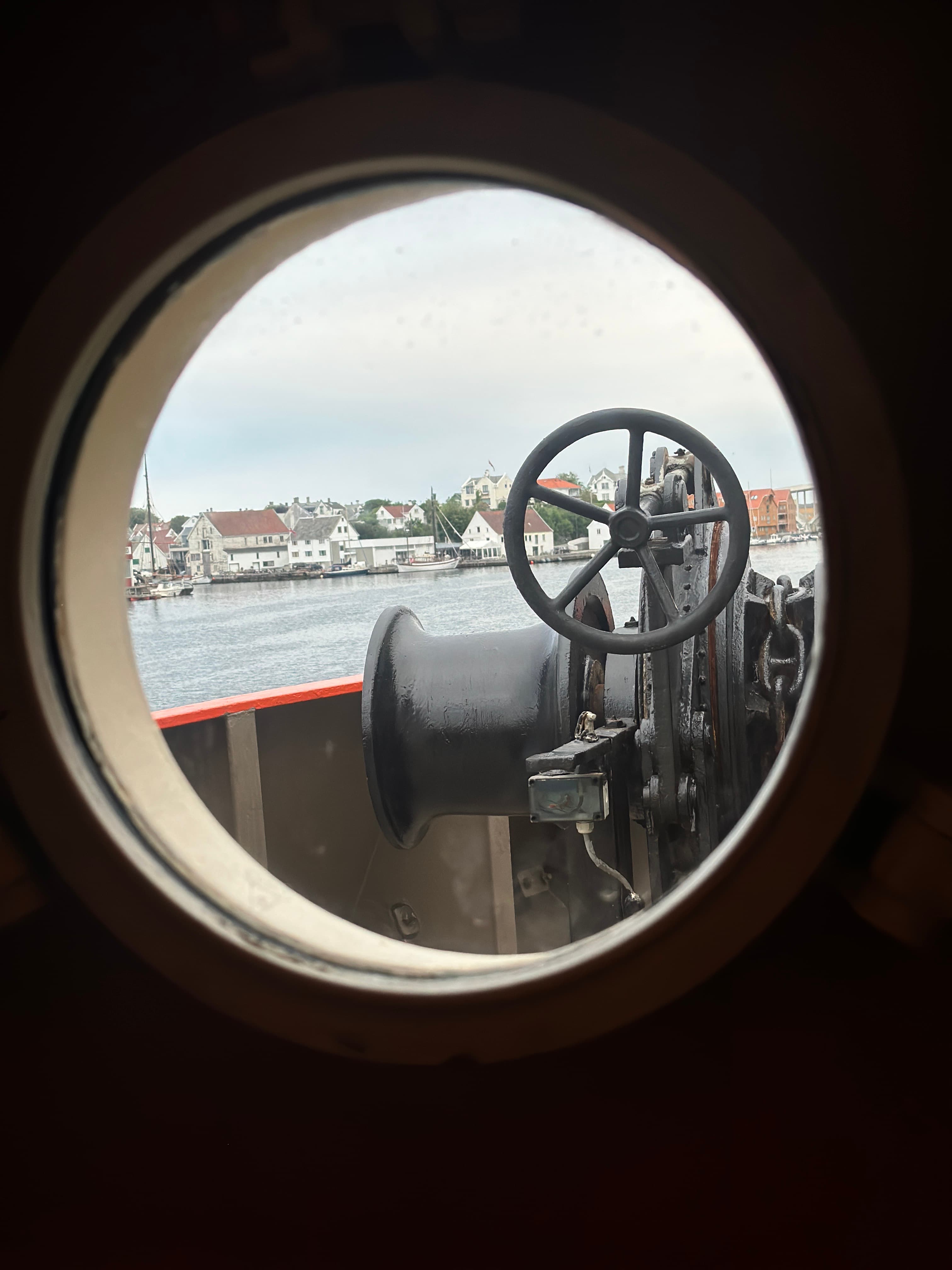 Circular porthole view of a black ship winch with a coastal town in the background.