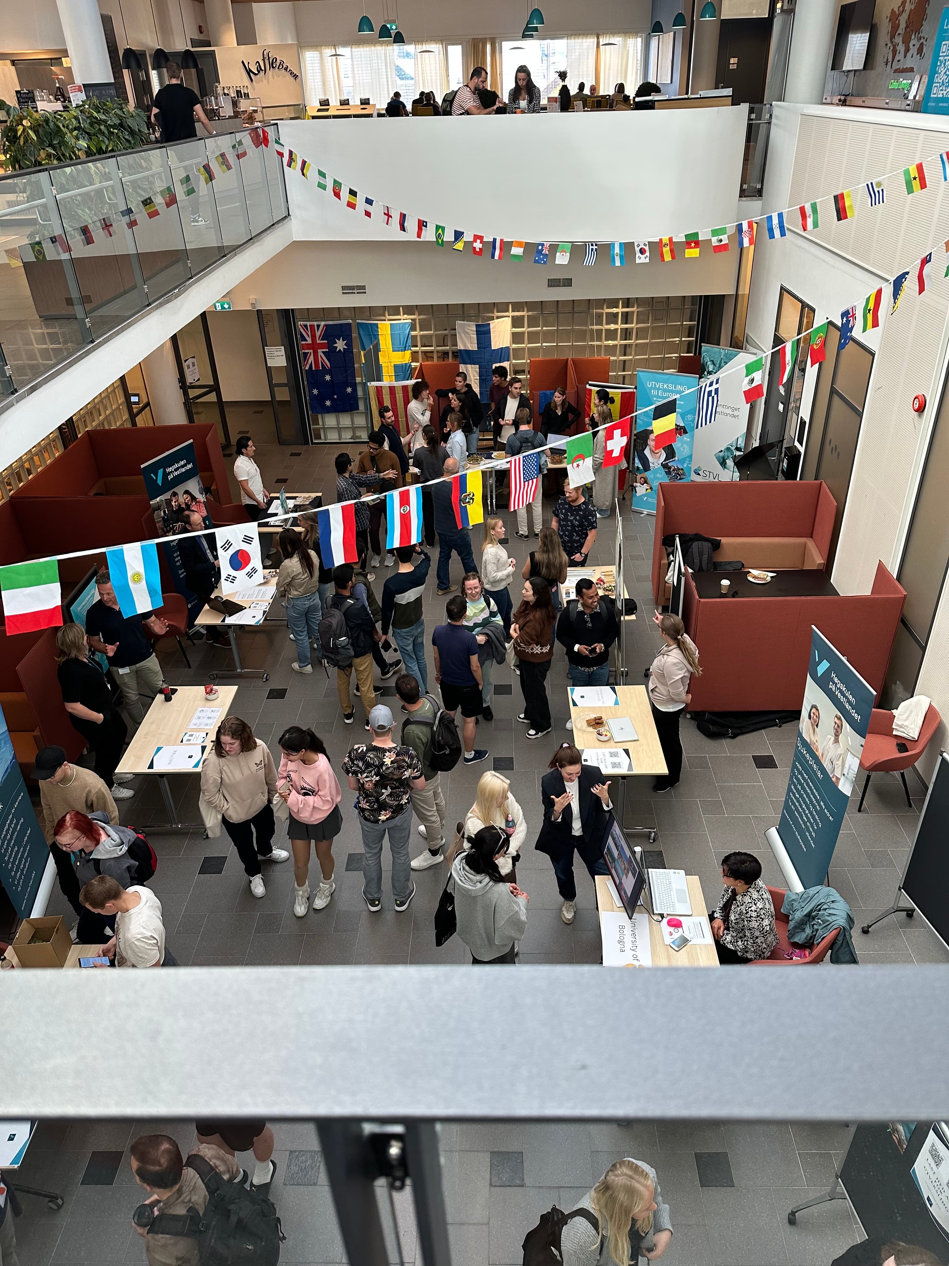 Crowded university atrium hosting an international fair with information booths and colorful flags overhead.