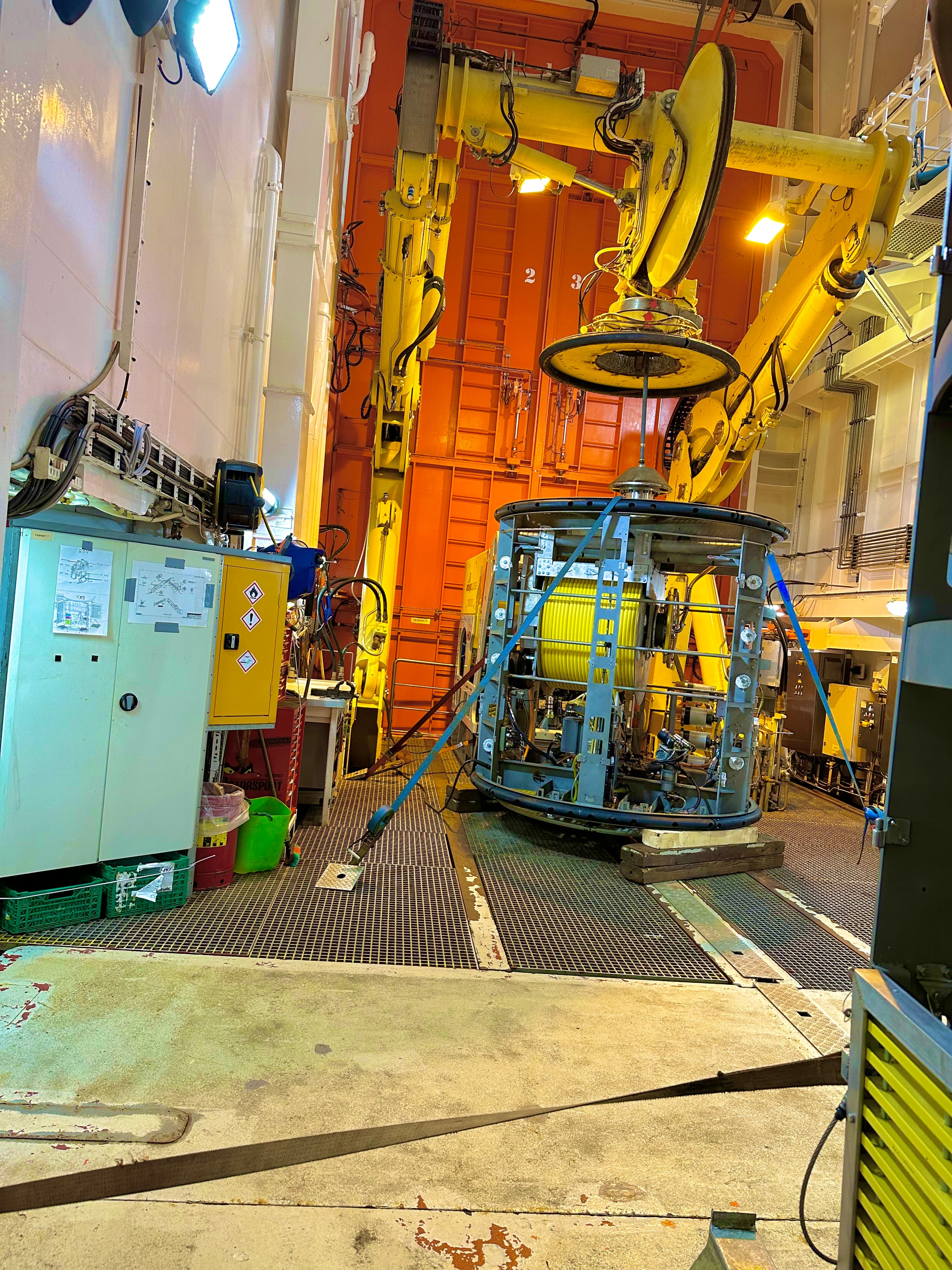 Yellow robotic arm over a cylindrical device with a yellow spool in an industrial hangar.