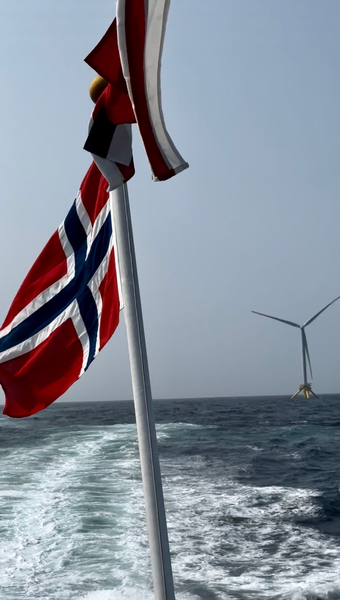 Norwegian flag on a boat's stern with a distant offshore wind turbine in the sea.