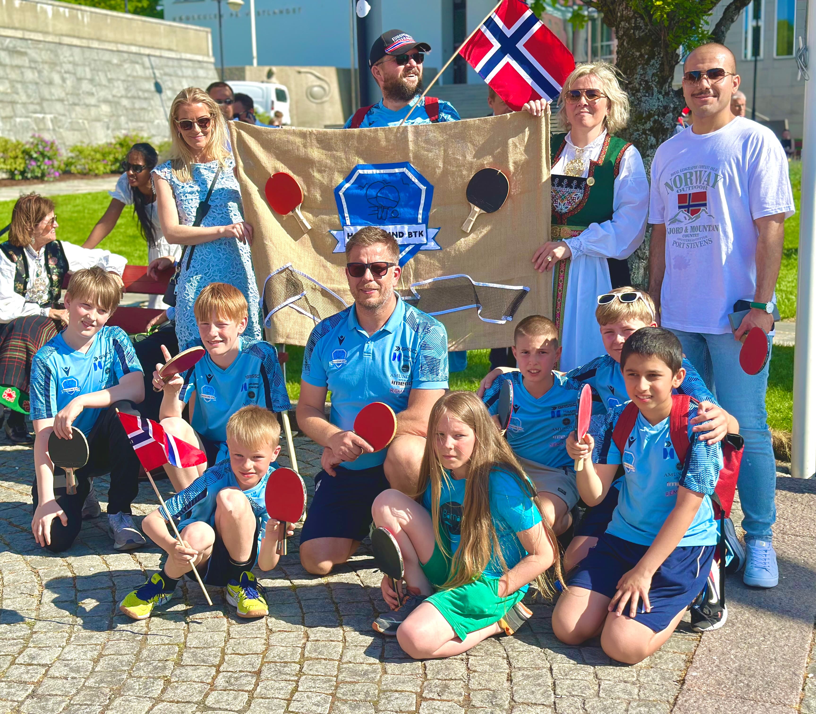 Norwegian table tennis club members pose with paddles and flags in front of a club banner.
