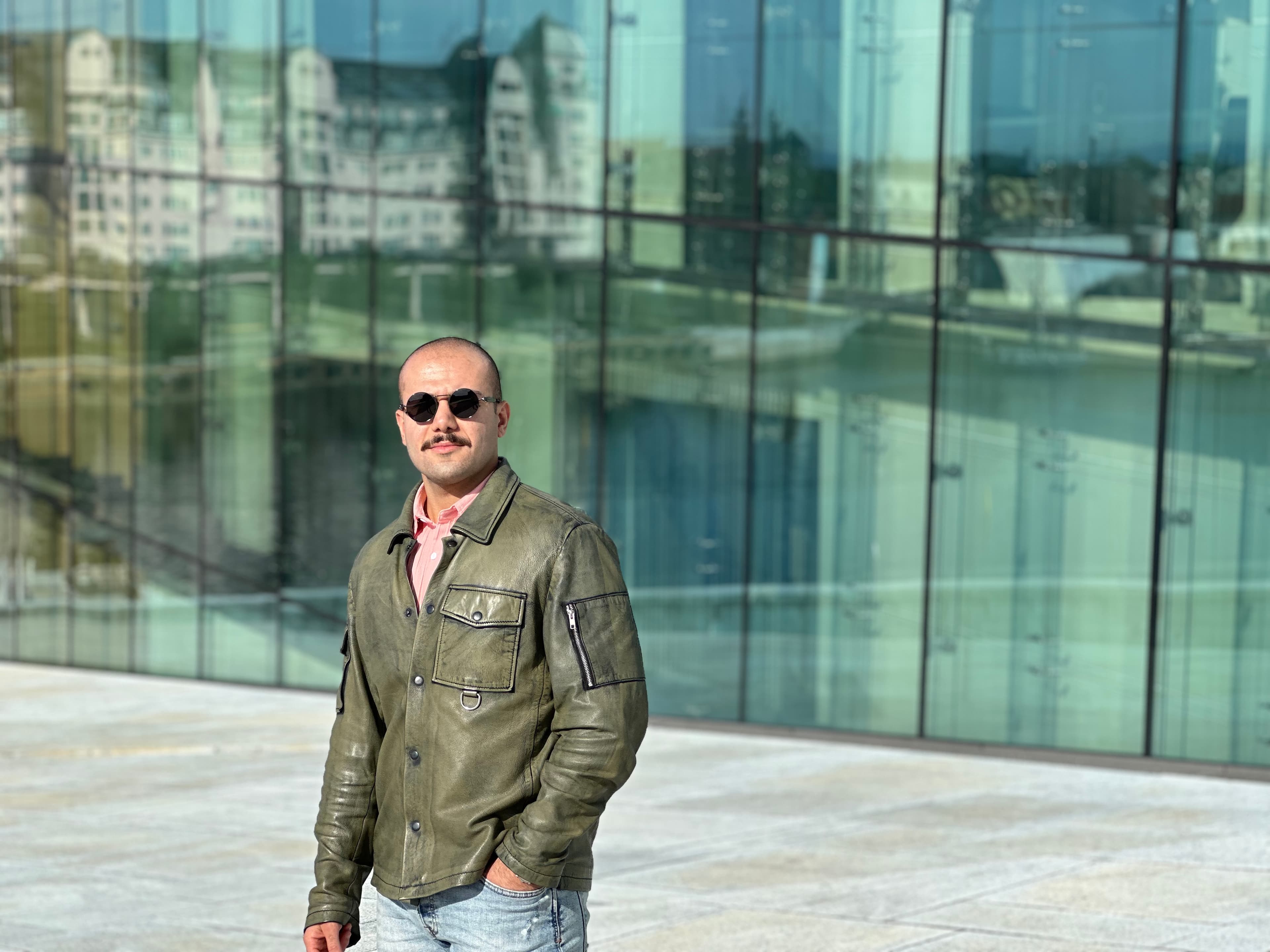 Man with mustache wearing green leather jacket and sunglasses in front of glass building.