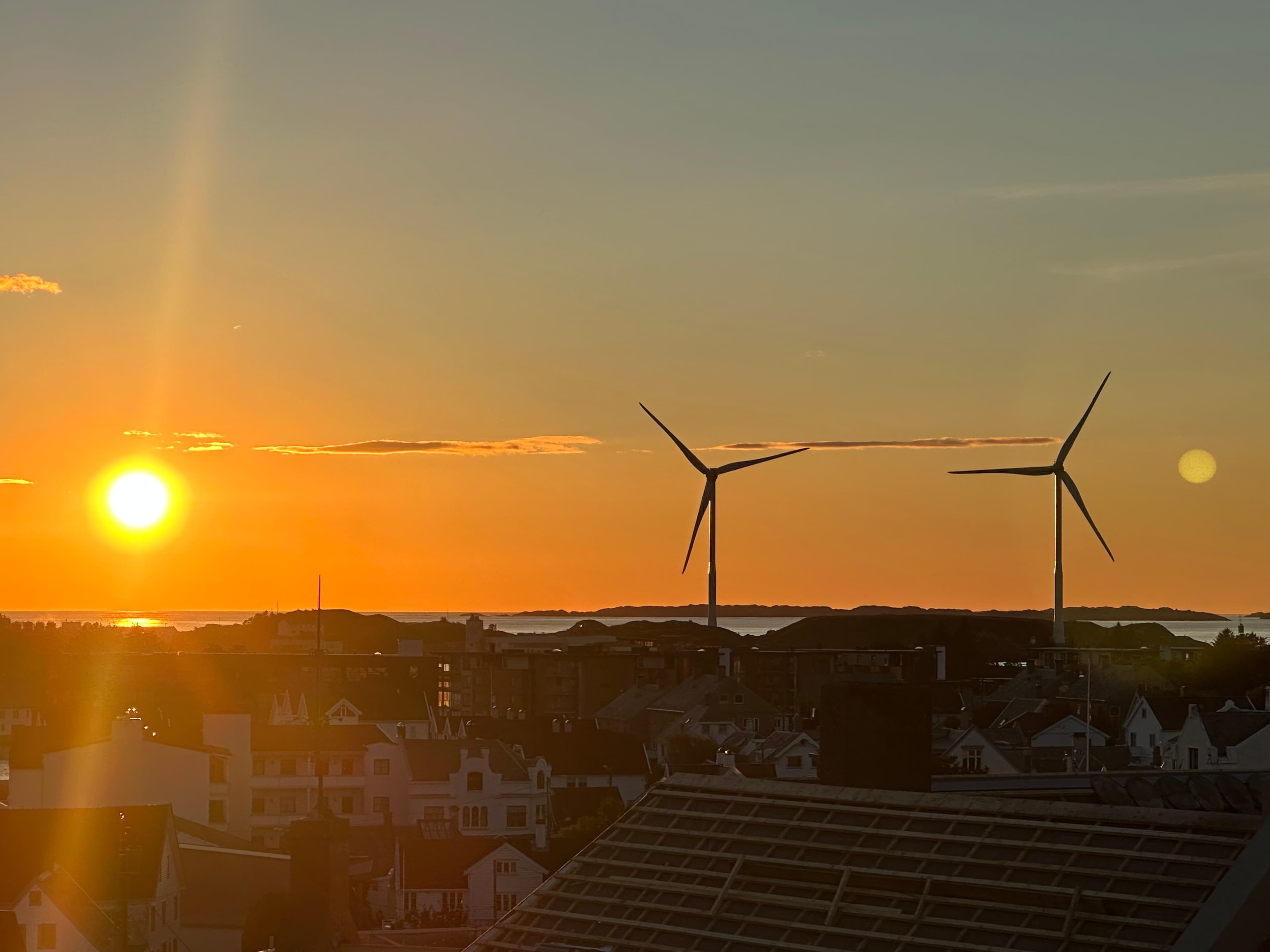 Two wind turbines silhouetted against a bright orange sunset over a coastal town and sea.