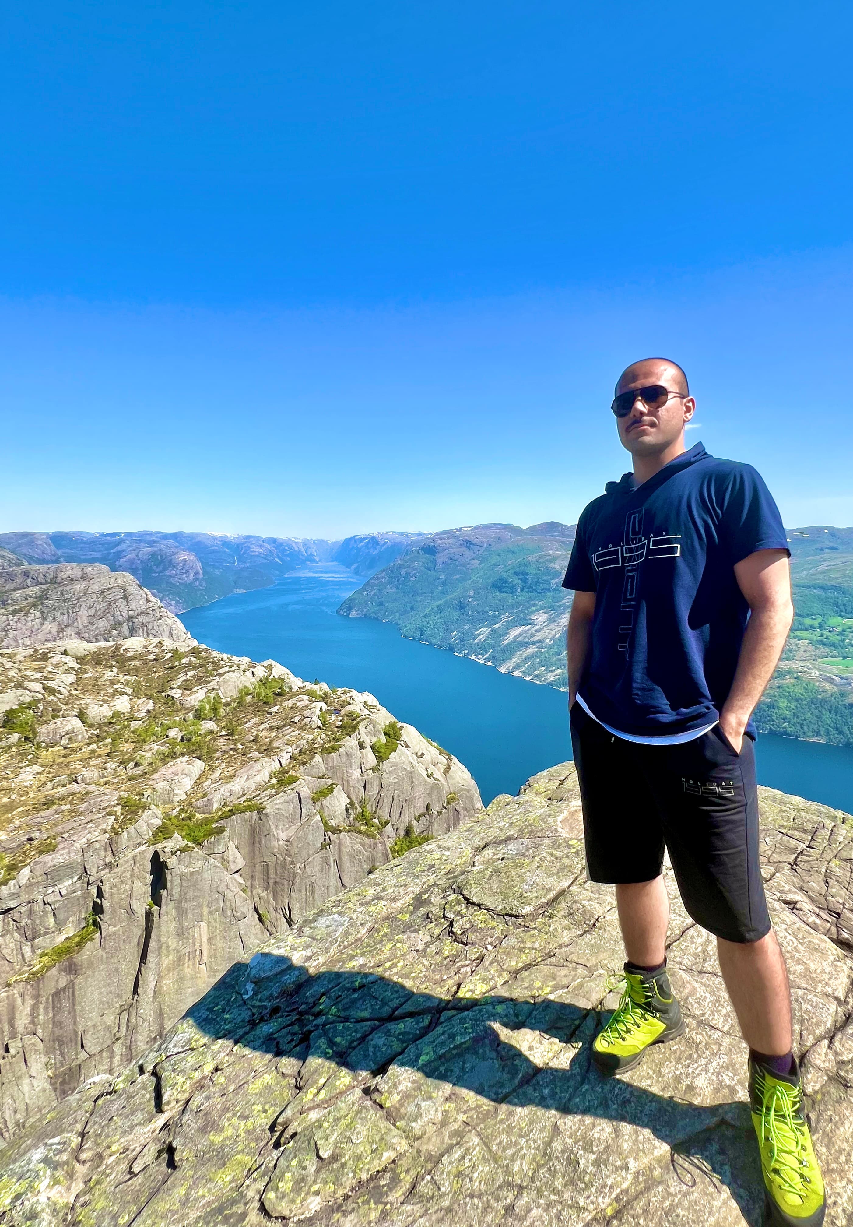 Man in green hiking boots stands on a rocky cliff overlooking a vast blue fjord.